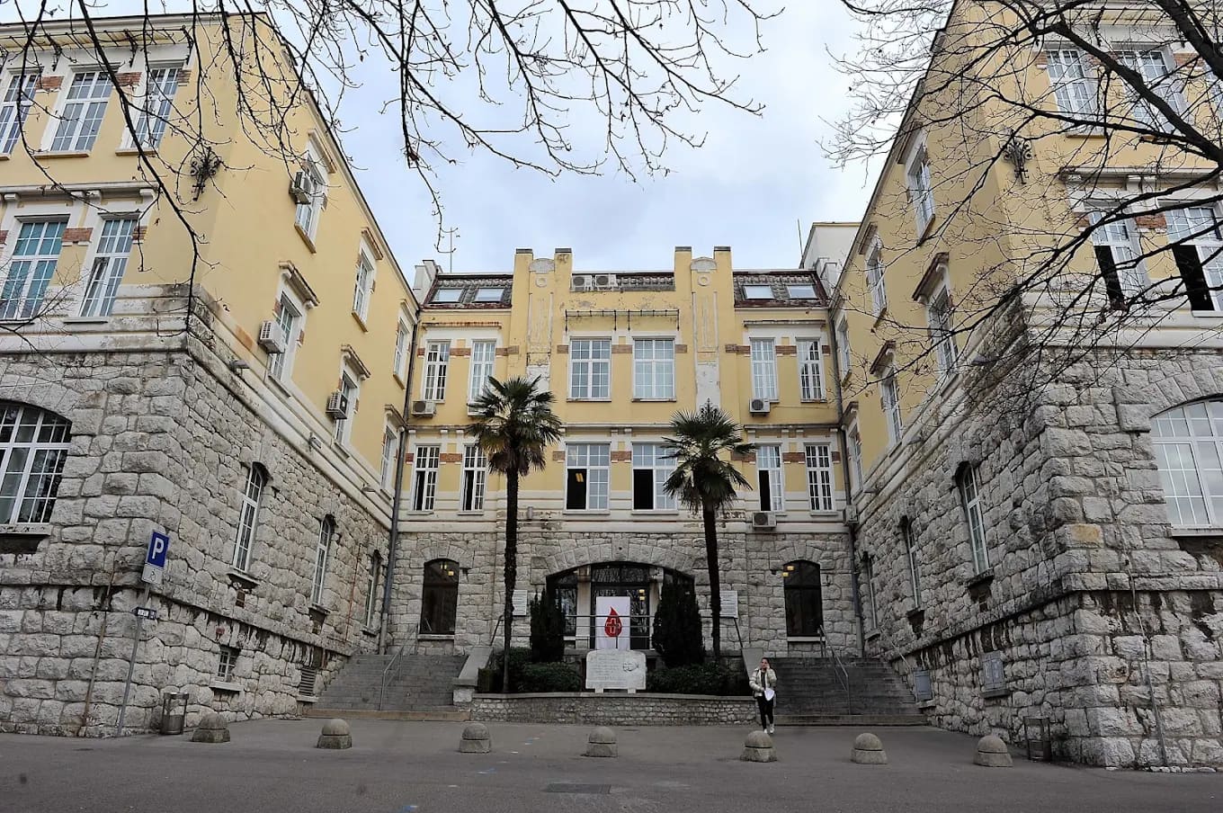 A historic yellow University of Rijeka faculty building with palm trees and a memorial in its courtyard, Rijeka, Croatia